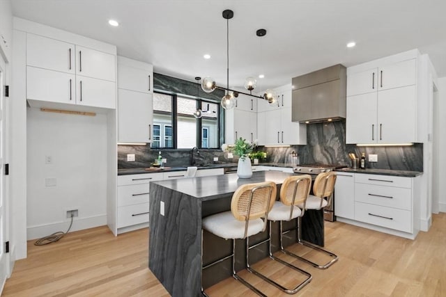 kitchen with a center island, hanging light fixtures, stainless steel stove, white cabinets, and wall chimney range hood