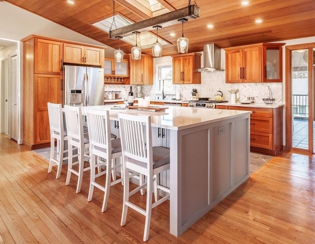 kitchen with stainless steel fridge with ice dispenser, wooden ceiling, wall chimney exhaust hood, a kitchen island, and light wood-style floors