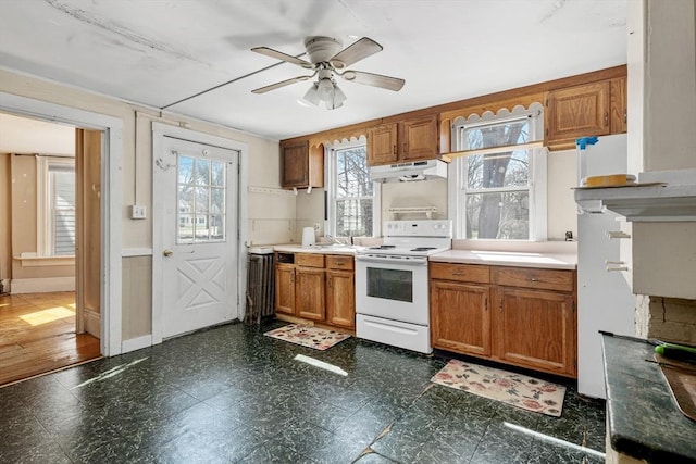 kitchen with white electric range oven, dark floors, a ceiling fan, brown cabinetry, and under cabinet range hood