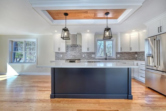 kitchen with high end refrigerator, wall chimney range hood, light countertops, and a tray ceiling