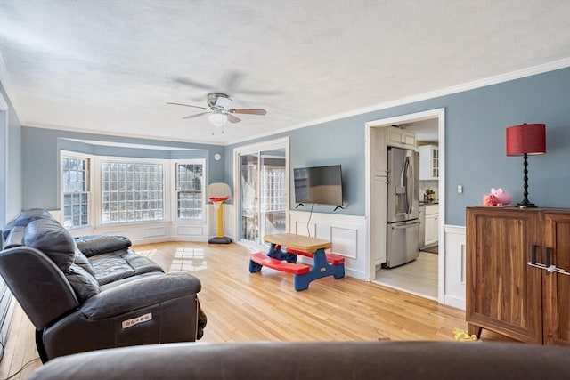 living area with light wood-style flooring, ornamental molding, ceiling fan, and wainscoting