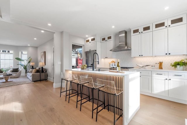 kitchen with a kitchen island with sink, wall chimney range hood, light hardwood / wood-style flooring, white cabinetry, and a breakfast bar area