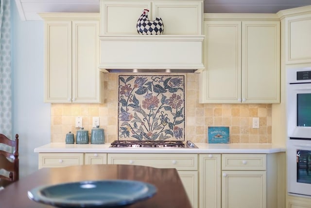 kitchen with white double oven, decorative backsplash, stainless steel gas stovetop, and cream cabinetry