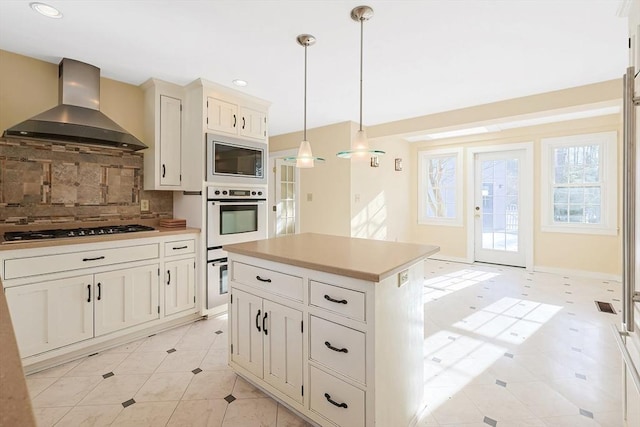 kitchen with stainless steel gas cooktop, wall chimney range hood, a center island, oven, and hanging light fixtures