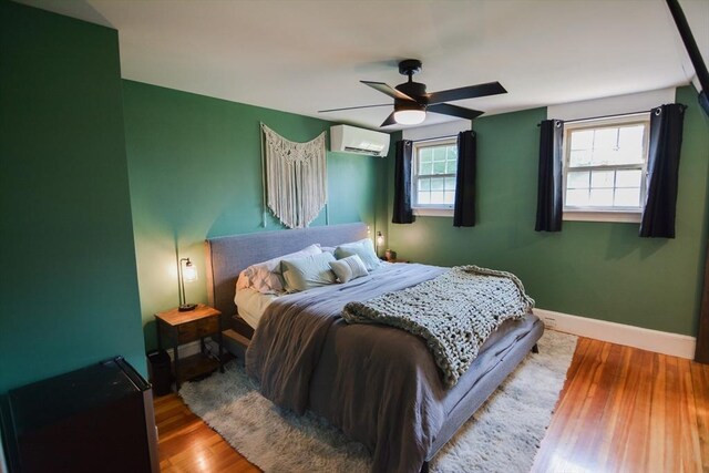 bedroom featuring a wall mounted AC, wood-type flooring, multiple windows, and ceiling fan