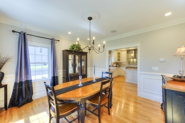 dining space featuring light wood-type flooring, a wainscoted wall, a notable chandelier, ornamental molding, and a decorative wall