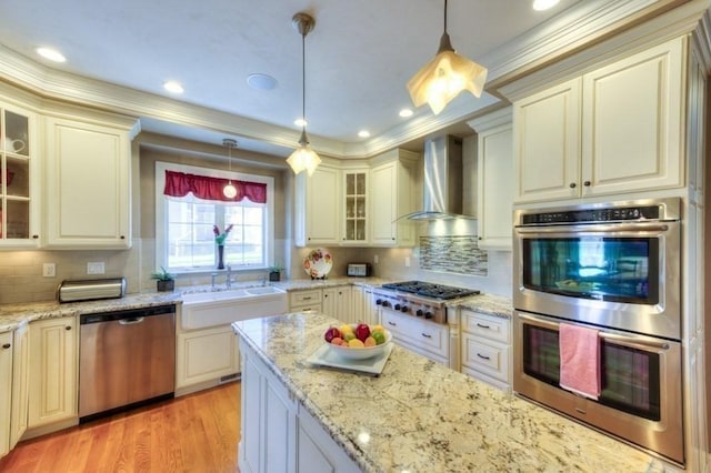 kitchen featuring a sink, wall chimney exhaust hood, stainless steel appliances, pendant lighting, and glass insert cabinets