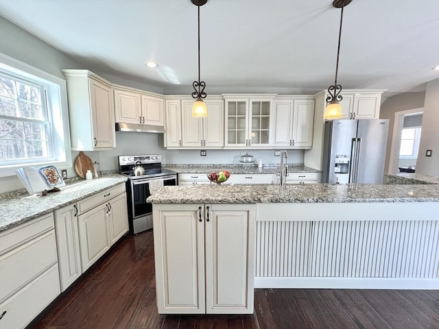 kitchen featuring stainless steel appliances, under cabinet range hood, dark wood-style flooring, pendant lighting, and an island with sink