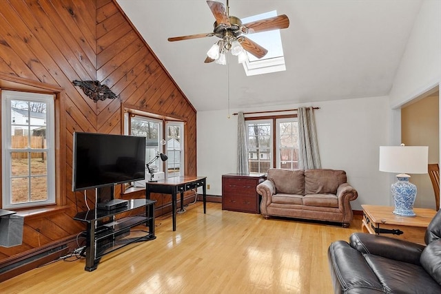 living area with high vaulted ceiling, light wood-style flooring, a skylight, ceiling fan, and wood walls