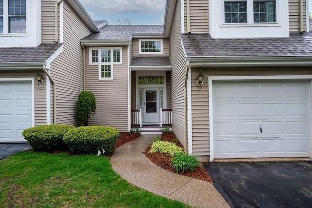 view of exterior entry featuring an attached garage, roof with shingles, and asphalt driveway