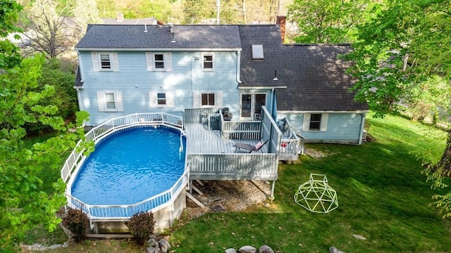 back of property with a deck, roof with shingles, an outdoor pool, a chimney, and a yard