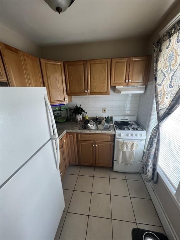 kitchen featuring brown cabinetry, white appliances, under cabinet range hood, backsplash, and light tile patterned floors