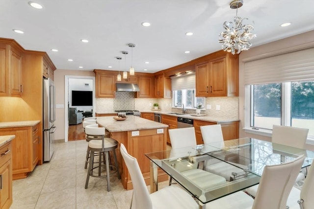 kitchen featuring a kitchen island, appliances with stainless steel finishes, decorative light fixtures, under cabinet range hood, and a sink