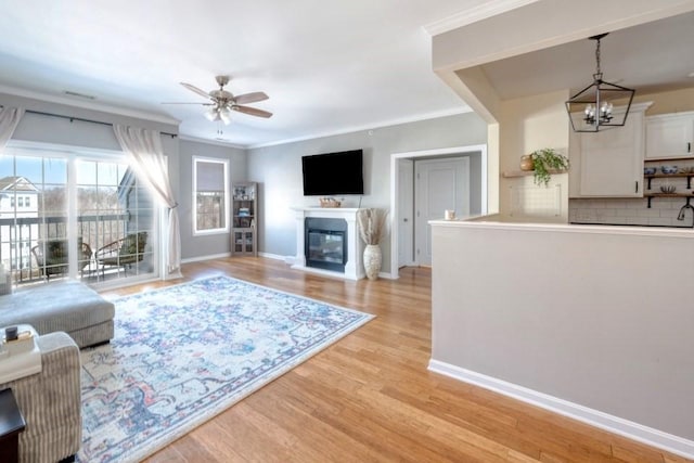 living room featuring crown molding, light wood-style flooring, a glass covered fireplace, baseboards, and ceiling fan with notable chandelier