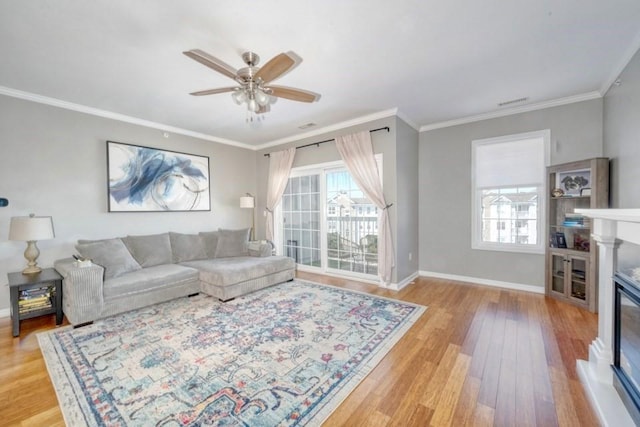 living area featuring light wood finished floors, baseboards, a ceiling fan, a glass covered fireplace, and crown molding