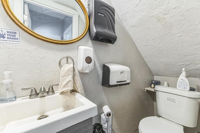 half bathroom featuring a textured wall, a sink, toilet, vaulted ceiling, and a textured ceiling