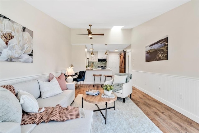 living room featuring light wood-style flooring, a barn door, a wainscoted wall, ceiling fan, and baseboards