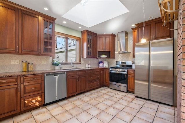 kitchen featuring wall chimney range hood, brown cabinets, a skylight, stainless steel appliances, and a sink