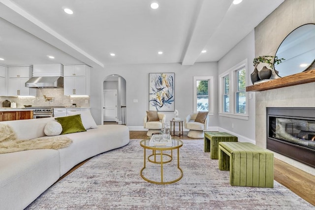 living area with arched walkways, wood finished floors, baseboards, beam ceiling, and a glass covered fireplace