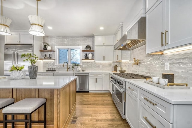 kitchen featuring open shelves, a sink, high quality appliances, light wood-type flooring, and wall chimney exhaust hood