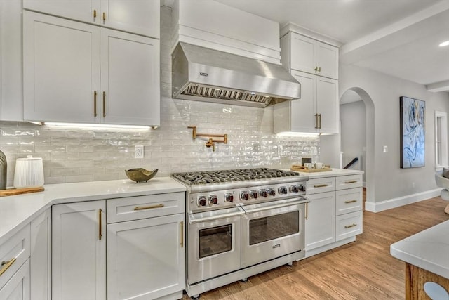 kitchen with range with two ovens, light countertops, light wood-style flooring, white cabinetry, and wall chimney range hood