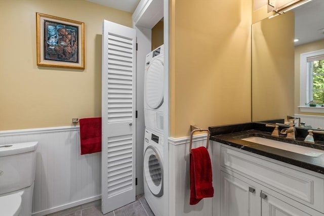 clothes washing area with a wainscoted wall, stacked washer and clothes dryer, a sink, tile patterned flooring, and laundry area