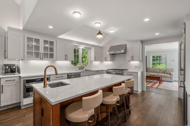 kitchen featuring a sink, a center island with sink, stainless steel appliances, wall chimney exhaust hood, and dark wood-style flooring