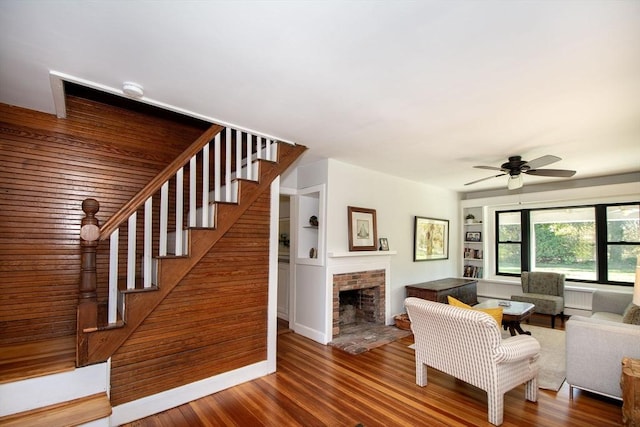 unfurnished living room featuring a brick fireplace, ceiling fan, built in shelves, and hardwood / wood-style floors