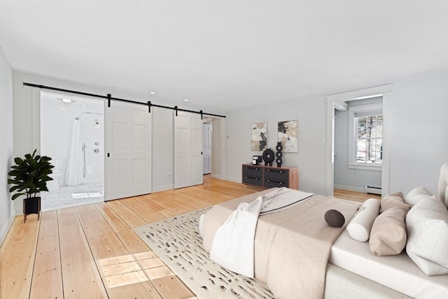 bedroom with baseboards, a barn door, light wood-style flooring, ensuite bathroom, and a baseboard radiator