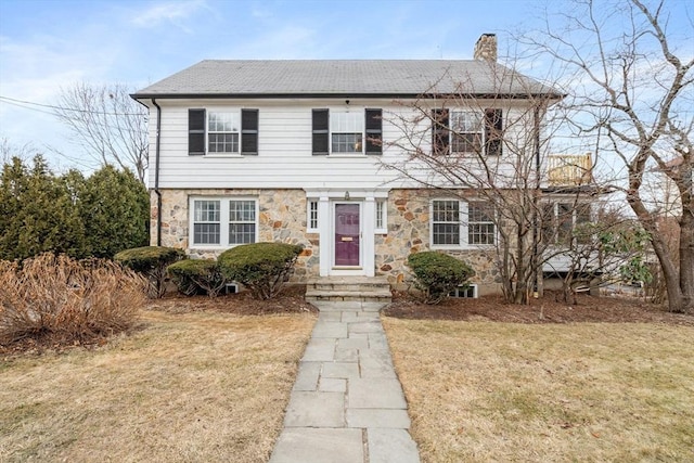 colonial inspired home featuring stone siding, a front yard, roof with shingles, and a chimney