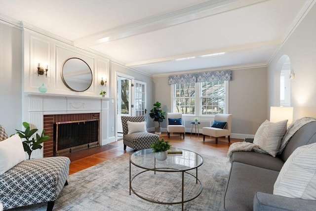 living area featuring beam ceiling, a brick fireplace, wood finished floors, and ornamental molding