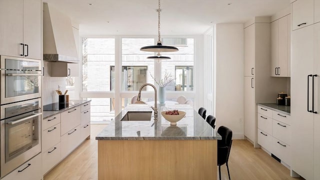 kitchen featuring wall chimney range hood, an island with sink, a sink, and modern cabinets
