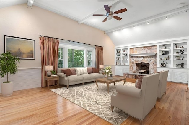 living area with a ceiling fan, lofted ceiling with beams, wood finished floors, a stone fireplace, and built in shelves