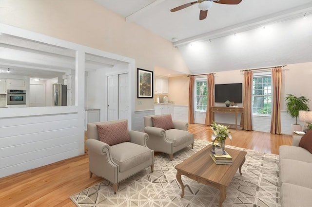 living area with lofted ceiling with beams, ceiling fan, light wood finished floors, and a wainscoted wall