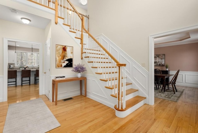 staircase with visible vents, a towering ceiling, wood finished floors, crown molding, and a decorative wall