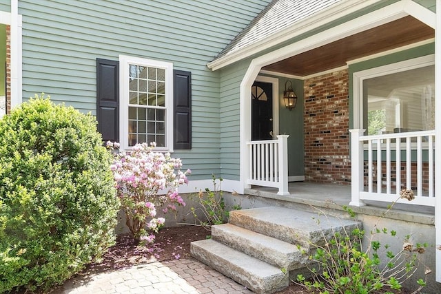 doorway to property featuring covered porch, brick siding, and roof with shingles