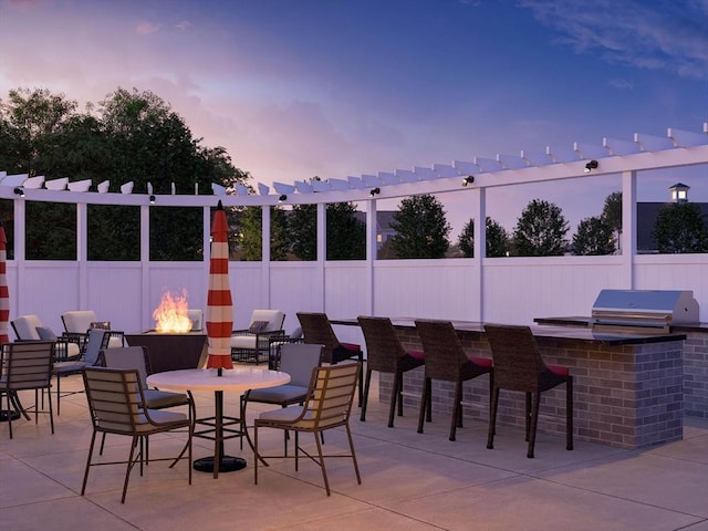 patio terrace at dusk featuring an outdoor fire pit, exterior kitchen, a grill, and fence