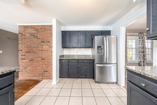 kitchen featuring light tile patterned flooring, light stone countertops, stainless steel fridge, brick wall, and backsplash