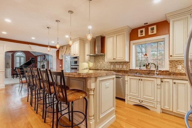 kitchen featuring wall chimney exhaust hood, light stone counters, cream cabinets, decorative light fixtures, and a kitchen island