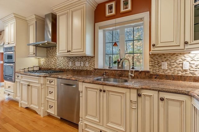 kitchen with sink, wall chimney exhaust hood, stainless steel appliances, dark stone counters, and cream cabinetry