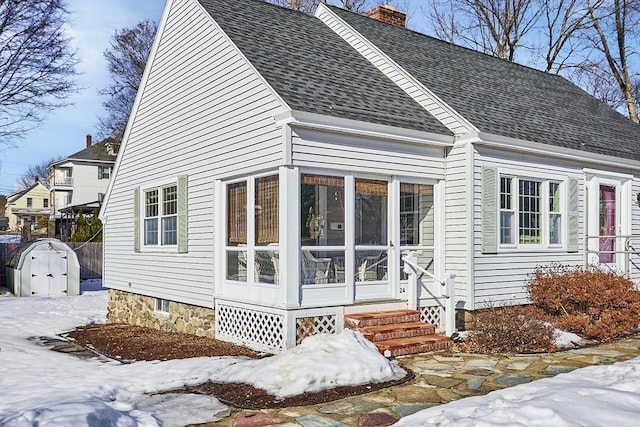 view of snowy exterior featuring a shingled roof, entry steps, a sunroom, and a chimney