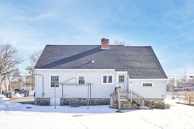 snow covered house with a shingled roof, fence, and a chimney