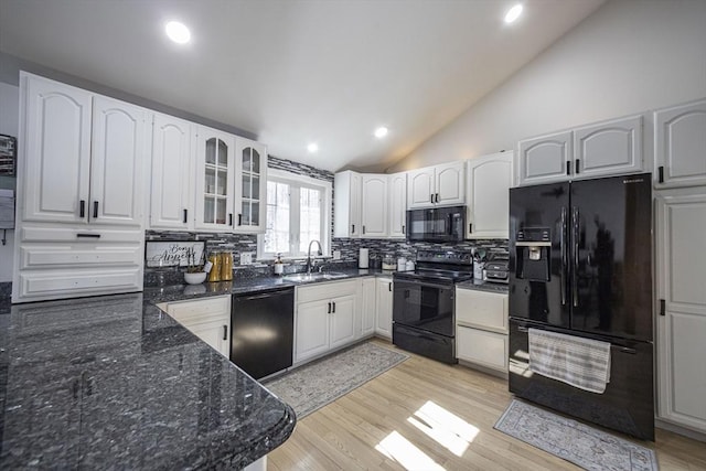 kitchen featuring lofted ceiling, a sink, black appliances, dark stone countertops, and glass insert cabinets