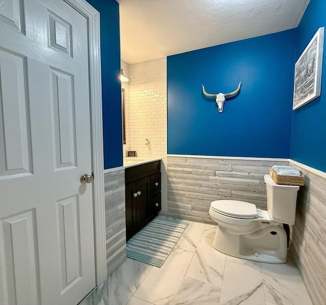 half bath with a wainscoted wall, toilet, marble finish floor, and vanity