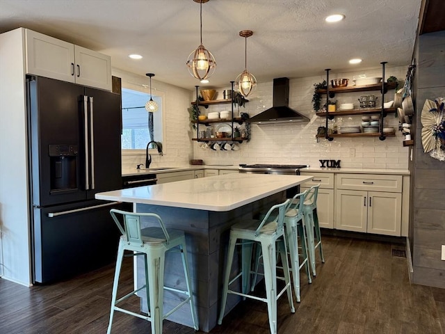 kitchen with open shelves, wall chimney range hood, dark wood finished floors, black refrigerator with ice dispenser, and a sink