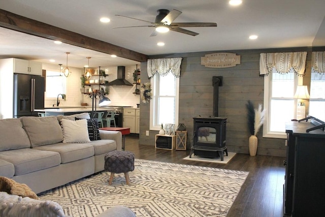 living room with beamed ceiling, dark wood-style flooring, a wood stove, and ceiling fan