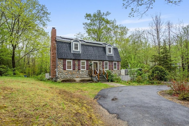 colonial inspired home featuring a chimney, stone siding, solar panels, driveway, and roof with shingles