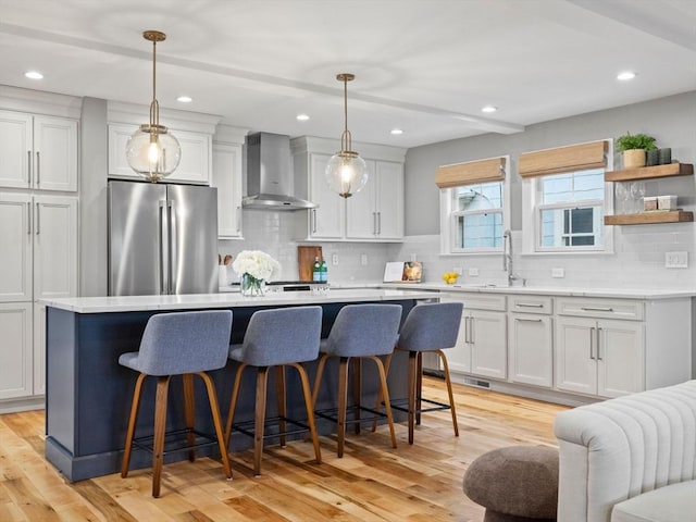 kitchen with stainless steel refrigerator, wall chimney exhaust hood, hanging light fixtures, and white cabinets