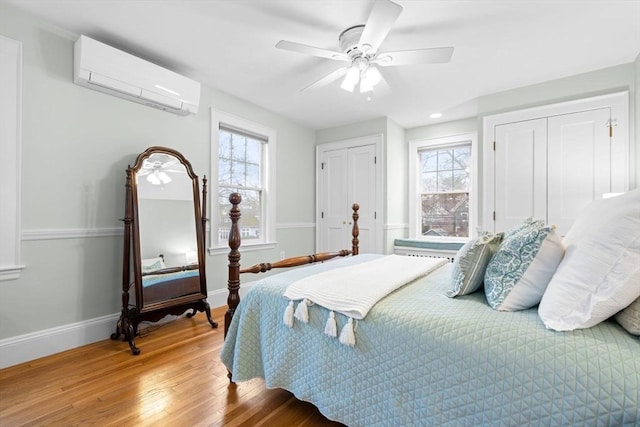 bedroom with multiple closets, light wood-style flooring, a wall mounted AC, ceiling fan, and baseboards