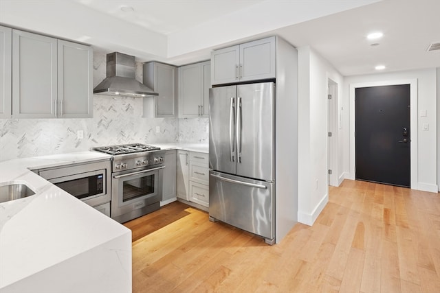 kitchen featuring appliances with stainless steel finishes, wall chimney exhaust hood, tasteful backsplash, and light hardwood / wood-style flooring